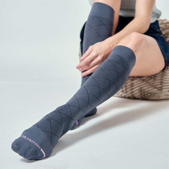 Knee-high compression socks in dark gray with a diamond pattern, worn by a woman sitting on a textured surface.