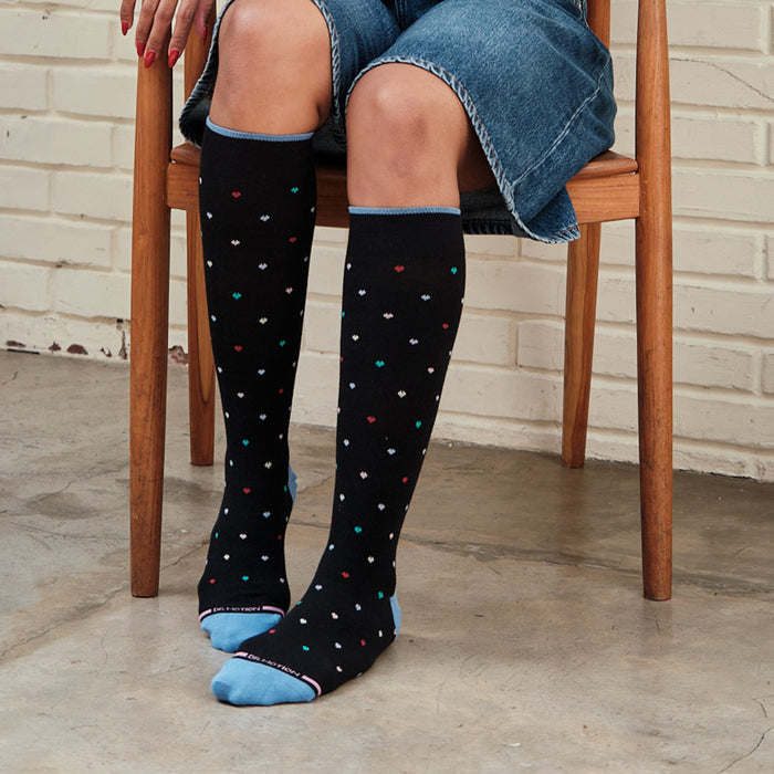 Knee-high black compression socks with colorful heart patterns and light blue cuffs, worn by a person sitting.