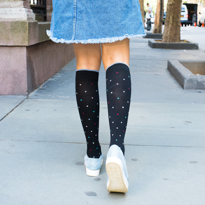 Knee-high black compression socks with colorful heart patterns, worn with a denim skirt and white shoes on a sidewalk.